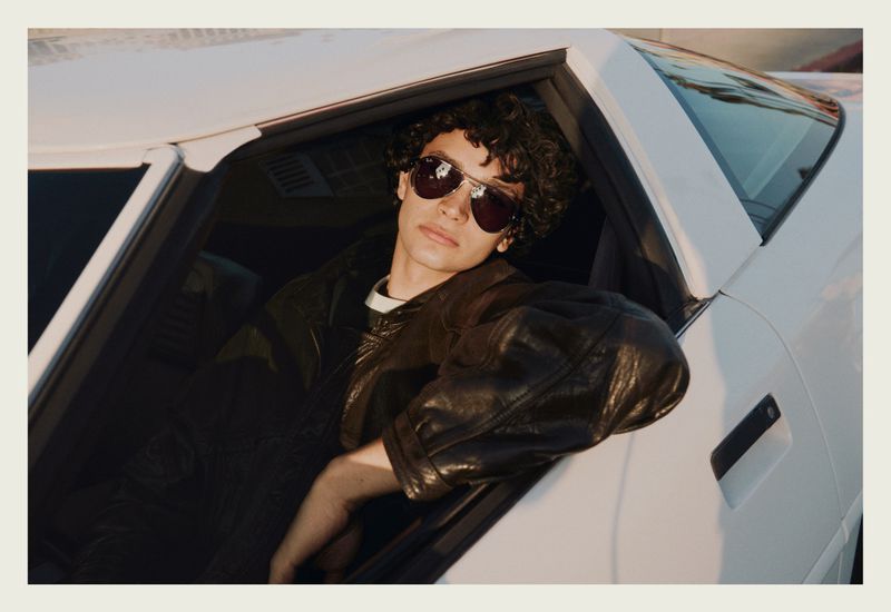A young man wearing aviator sunglasses and a black leather jacket leans out the window of a white car.
