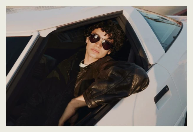 A young man wearing aviator sunglasses and a black leather jacket leans out the window of a white car.