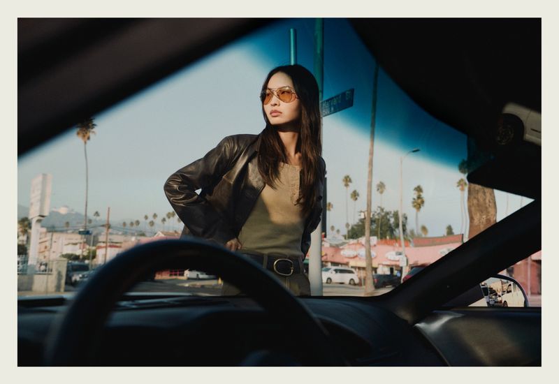 Model wearing aviator sunglasses and leather jacket standing outside viewed through a car windshield