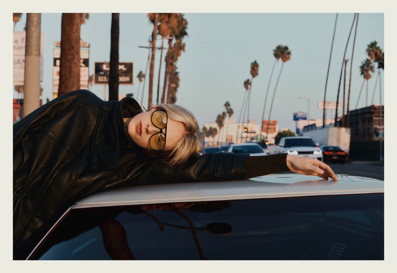 Model wearing aviator sunglasses and a black leather jacket leaning on a car roof under palm trees