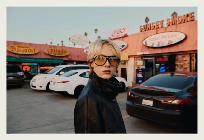 Blonde model wearing yellow-tinted aviator sunglasses and a black leather jacket in a shopping strip parking lot
