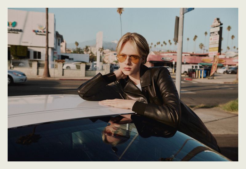Model wearing yellow sunglasses and black leather jacket leaning on white car hood on sunny street