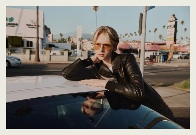 Model wearing yellow sunglasses and black leather jacket leaning on white car hood on sunny street