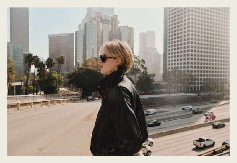 Model wearing dark sunglasses and a black leather jacket overlooking a multi-lane freeway and city skyline.