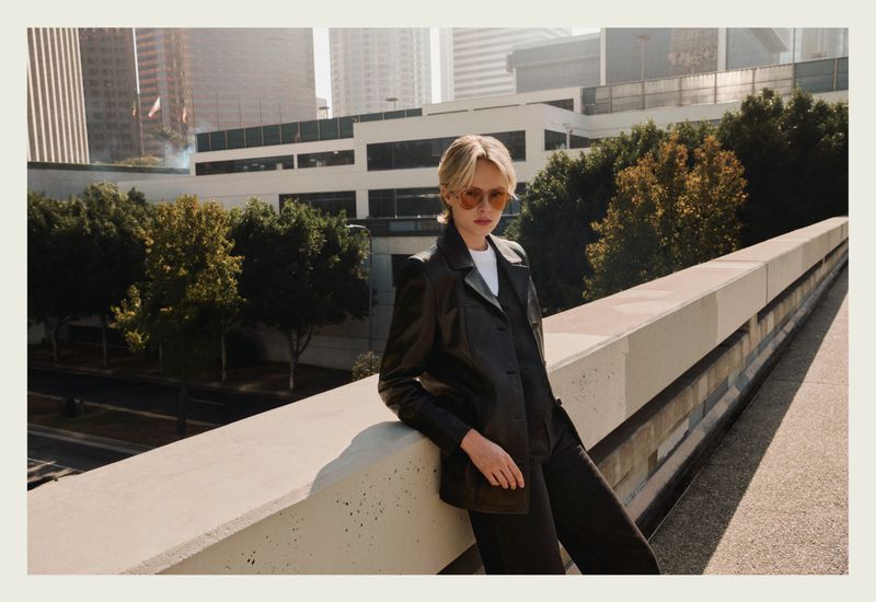 Model wearing tinted aviator sunglasses and a black leather jacket leaning on a concrete ledge against an urban backdrop.
