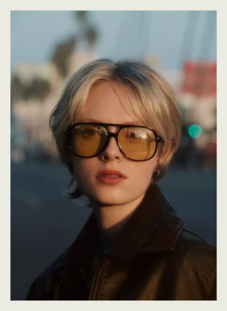 Model wearing amber-tinted aviator sunglasses and a dark jacket outdoors against a softly blurred city street background.