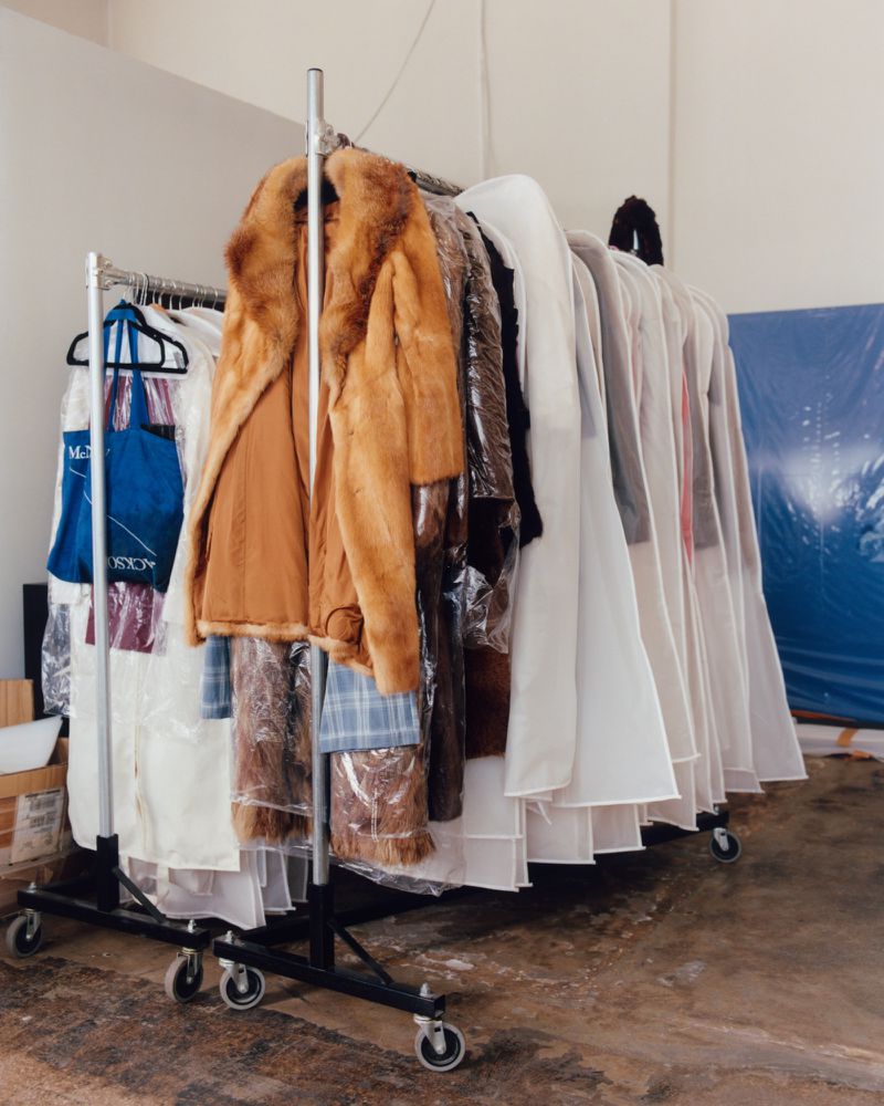 Rolling racks filled with garment bags and clothing in a studio space with a prominent brown fur coat hung on the front rail.