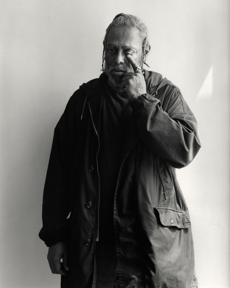 Portrait of artist Arthur Jafa touching his eye while wearing a dark jacket against a plain light wall background.
