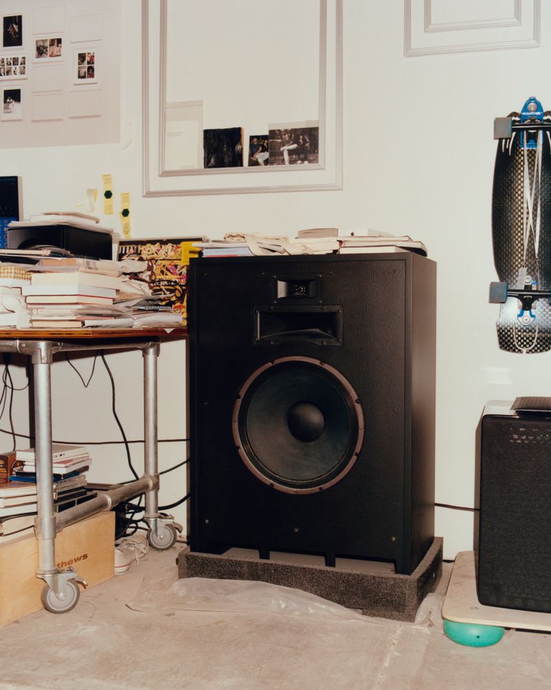 Large black speaker cabinet resting on a foam base inside a room with stacked books, a metal rolling table, and skateboard on the wall
