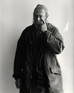 Portrait of artist Arthur Jafa touching his eye while wearing a dark jacket against a plain light wall background.