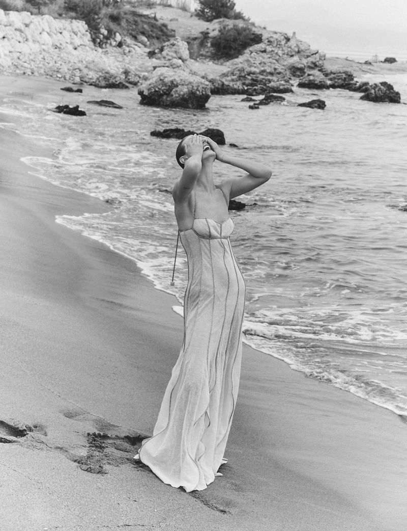 Model Tanya Ruban standing on a sandy beach wearing a long light-colored dress with hands over her face