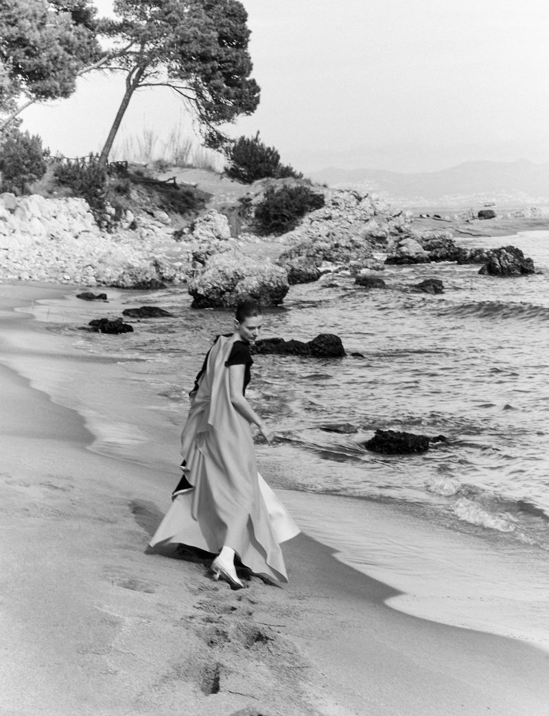 Model wearing a long flowing dress walking along a sandy beach near rocky coastlines and calm ocean waters