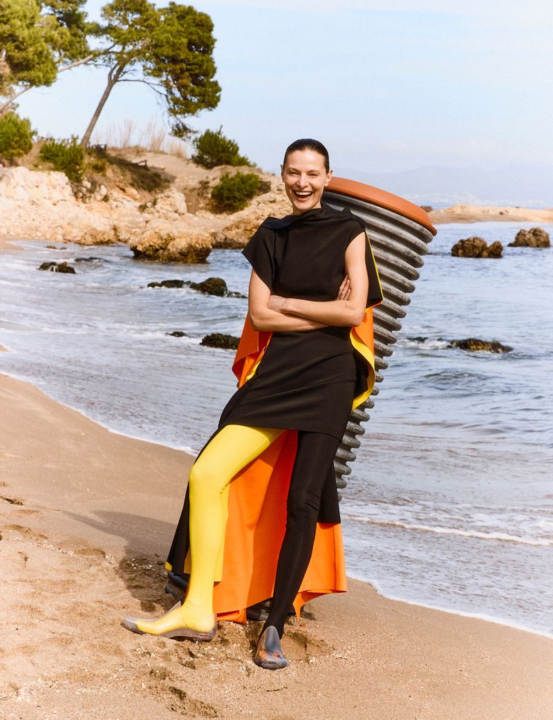 Model standing on a sandy beach wearing a black dress, orange cape, and one yellow legging for Flair Germany.