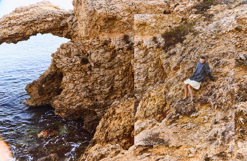 Model seated on a rocky coastal cliff next to the sea, captured in a composite framing style by photographers Andoni & Arantxa.