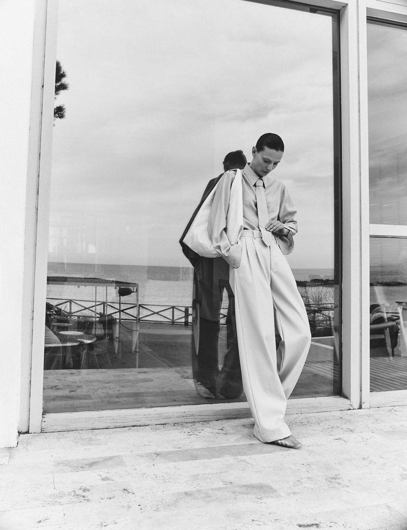 Model Tanya Ruban leaning against a large glass window wearing wide-leg trousers, a button-up shirt, and a tie in a black-and-white editorial