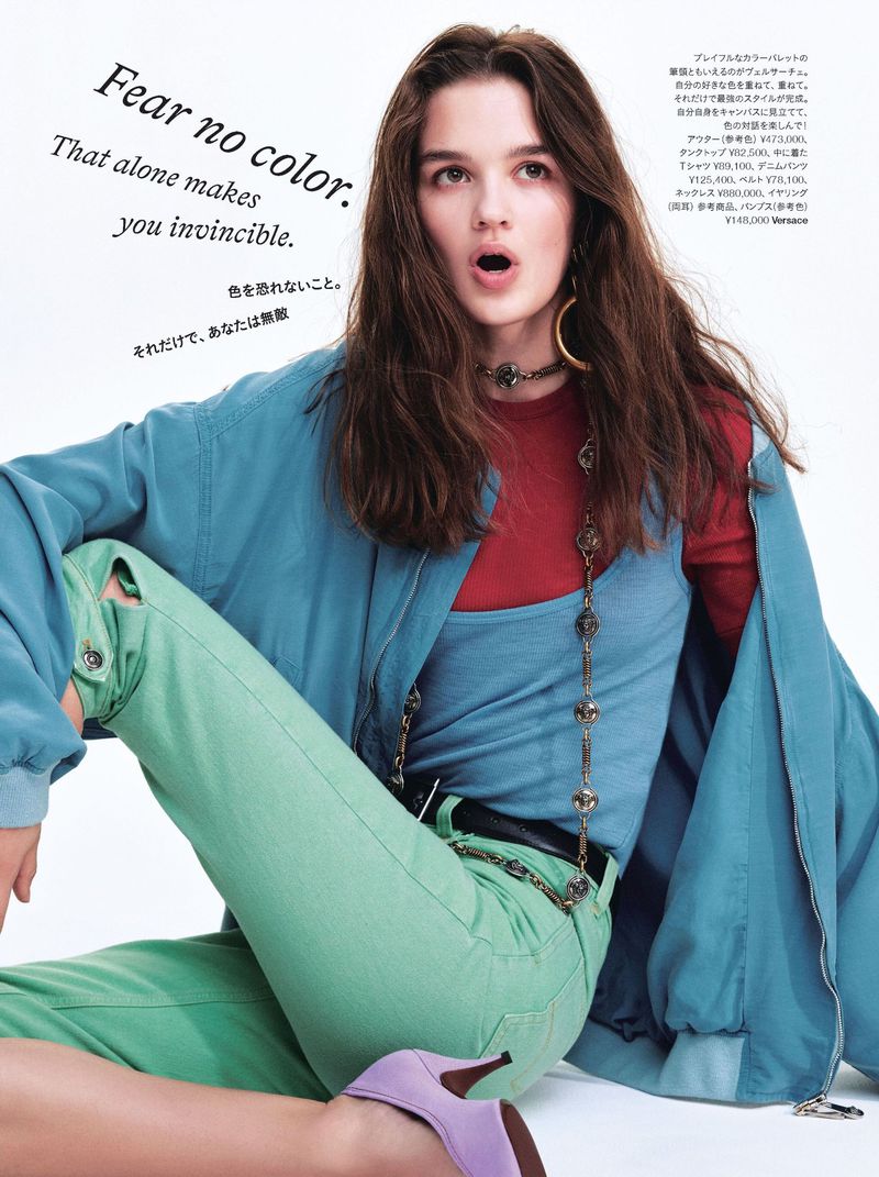 Model with long brown hair wearing a blue jacket, red top, green pants, and a purple shoe against a white background.