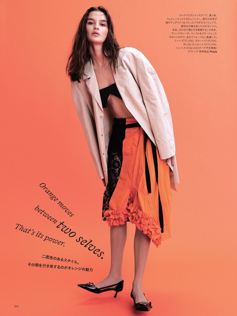 Fashion editorial shot of a woman in an orange skirt and light-beige jacket posing against an orange studio background with dark text.