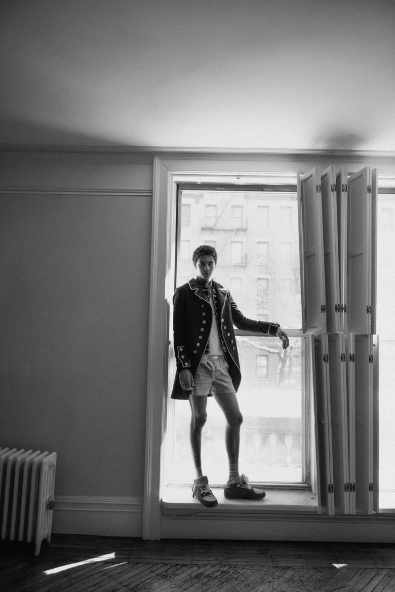 Monochrome portrait of a male model standing in an open window frame wearing a tailored jacket and shorts.