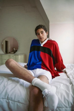 Young man seated on a white bed wearing a red, blue, and black rugby shirt with white shorts and socks.