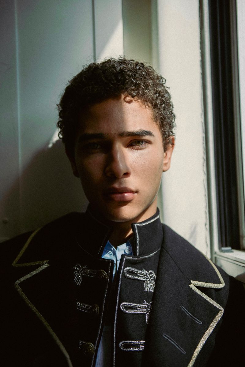 Close-up portrait of a young man with curly hair wearing a dark jacket with light contrast embroidery standing near a window