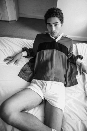 Young male model reclining on a white bed wearing a patterned rugby shirt and light-colored shorts.