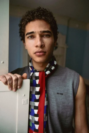 Close-up portrait of a young man with curly hair wearing a grey sleeveless top and a patterned scarf, holding a white doorframe.