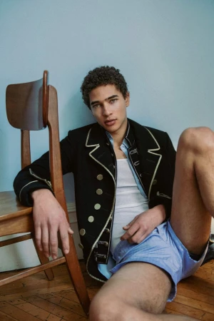Male model wearing a black military-style jacket and light blue shorts seated on a wooden floor leaning against a chair.