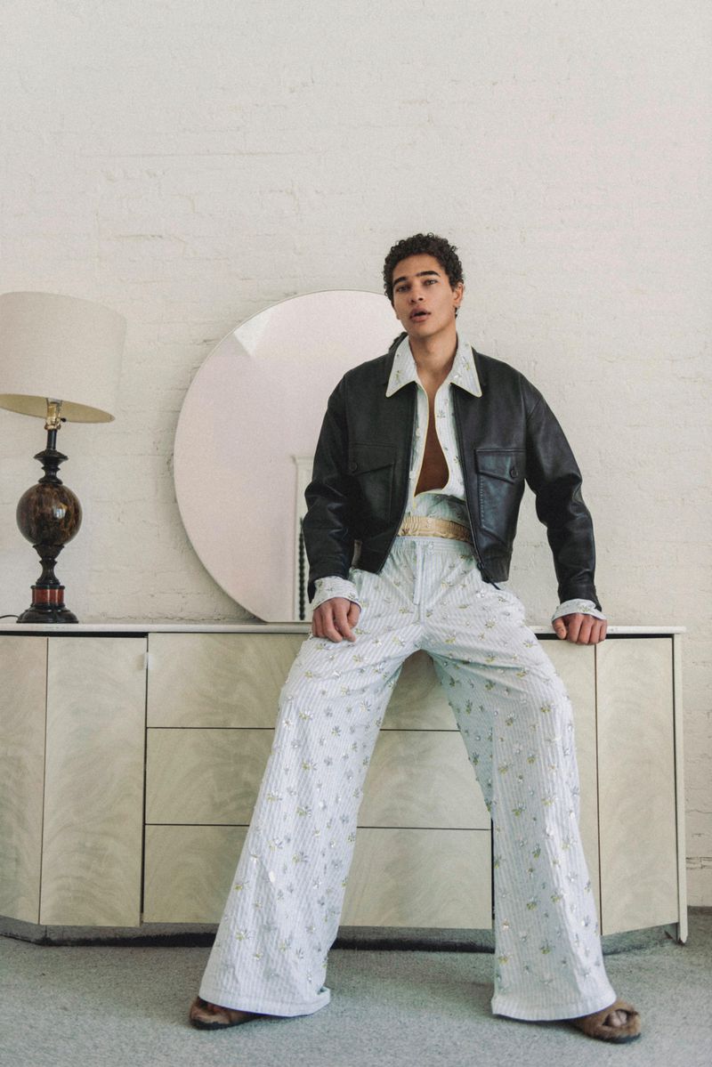 Model wearing a black leather jacket and patterned trousers seated on a white credenza with a round mirror and lamp.