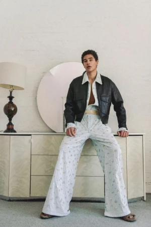 Model wearing a black leather jacket and patterned trousers seated on a white credenza with a round mirror and lamp.