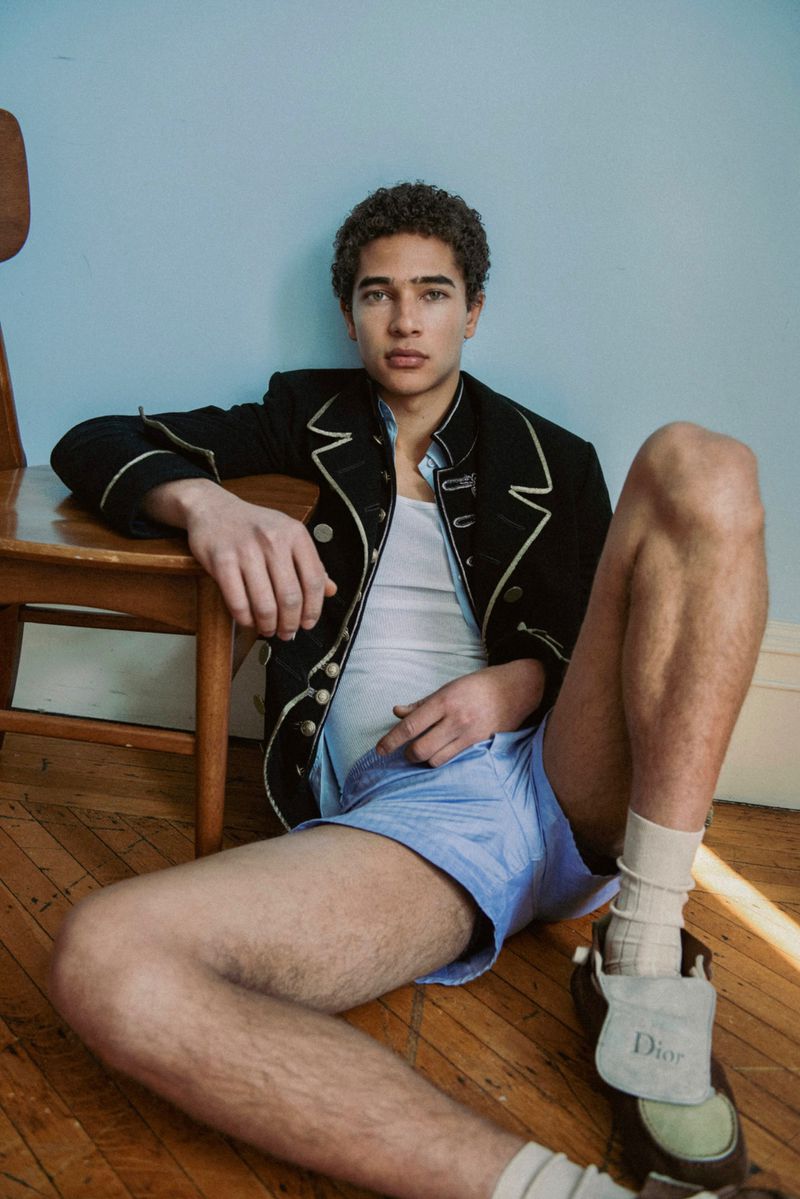 A young male model seated on a wooden floor leaning against a chair wearing a black jacket and blue shorts against a light blue wall.