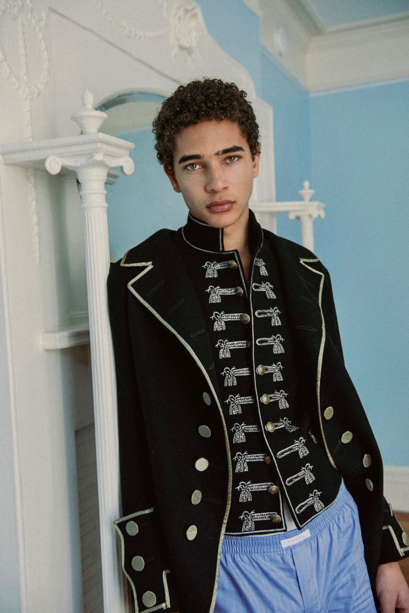 Young man with curly hair wearing an embroidered black military jacket and blue trousers against a pale blue wall.