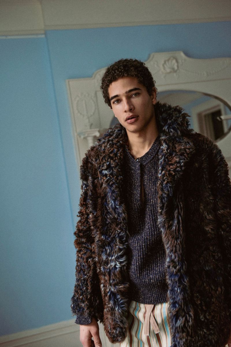 Model wearing a textured brown and blue faux fur coat against a pale blue wall and ornate architectural mirror frame.