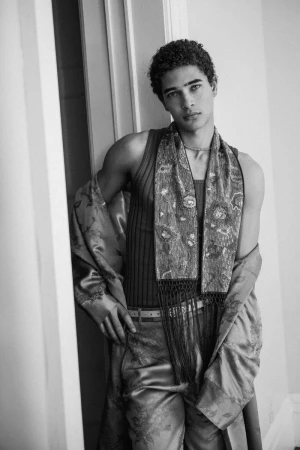 Monochrome portrait of a young male model leaning against a doorway wearing patterned trousers and a matching draped robe over a ribbed tank top