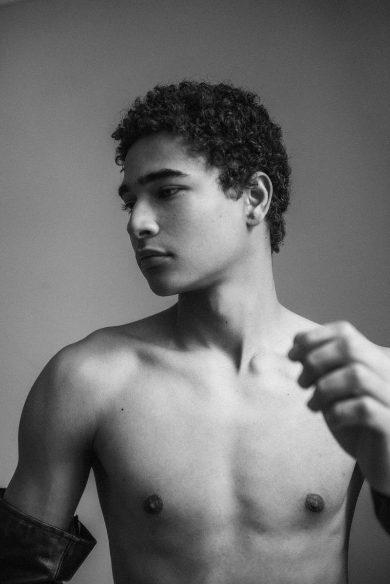 Monochrome portrait of a young male model with short curly hair looking thoughtfully to the side against a seamless grey backdrop.
