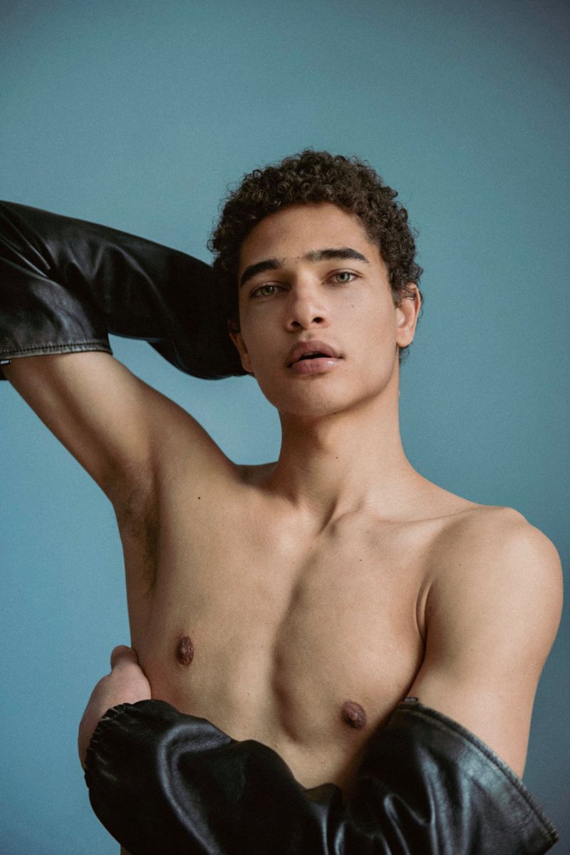 Close-up portrait of a shirtless young man with curly dark hair wearing leather sleeves against a blue background.