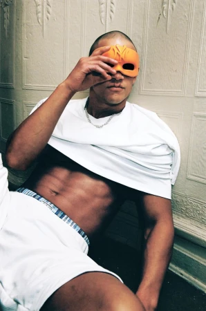 Model holding an orange mask over his eyes, wearing a white t-shirt and shorts against a paneled wall.