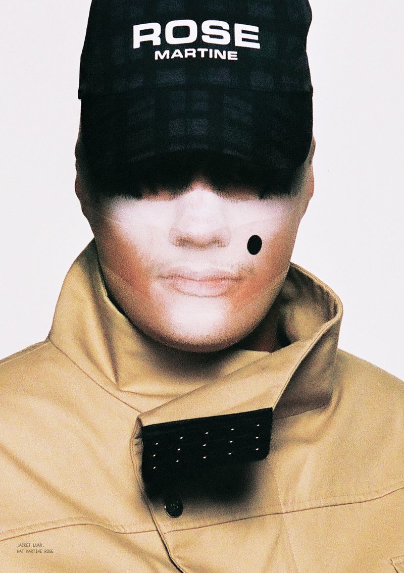 Close-up portrait of model wearing a black baseball cap with white text and a tan high-collared jacket against a white background
