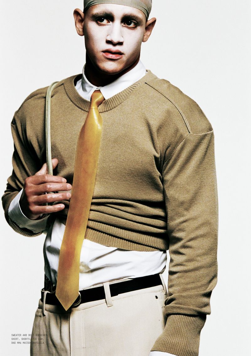 Model wearing a tan crewneck sweater over a white shirt with a yellow tie against a plain white background