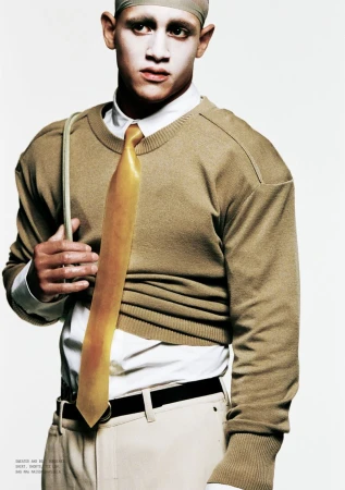 Model wearing a tan crewneck sweater over a white shirt with a yellow tie against a plain white background