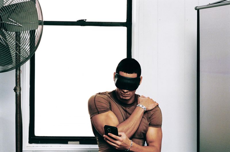 Man wearing a brown t-shirt and black blindfold holding a smartphone in front of a window and studio fan.