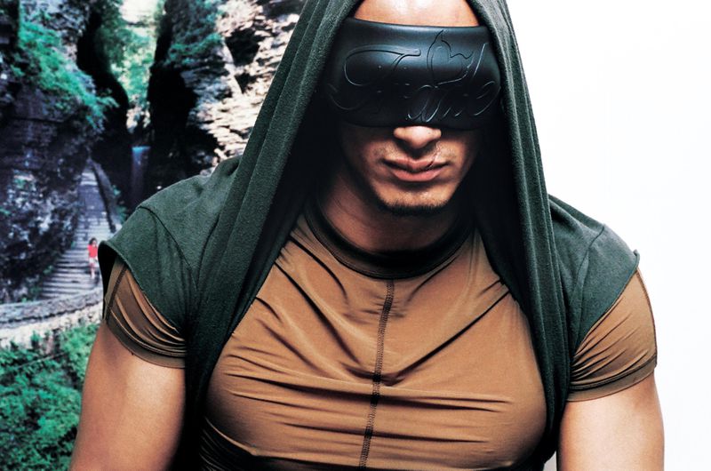 Man wearing a dark green hooded top and tan t-shirt with a black sculptural eye mask, photographed by Aidan Zamiri for i-D Magazine.