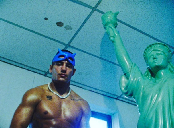 Shirtless man wearing a blue mask next to a green Statue of Liberty replica under a tiled ceiling