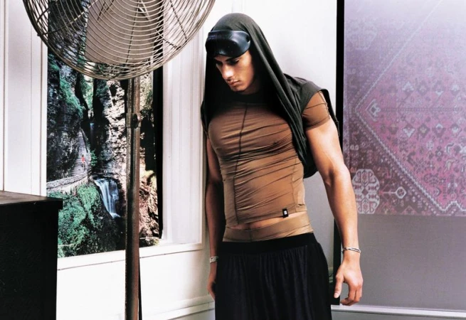 Model in brown fitted top and black trousers standing indoors next to a large floor fan with framed art on the wall