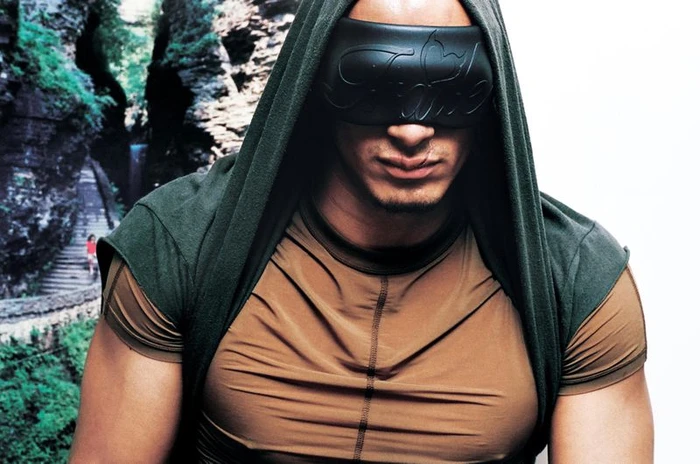 Man wearing a dark green hooded top and tan t-shirt with a black sculptural eye mask, photographed by Aidan Zamiri for i-D Magazine.