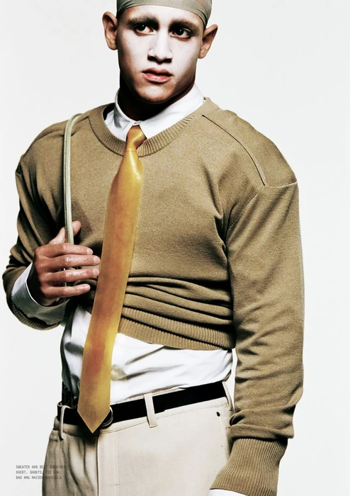 Model wearing a tan crewneck sweater over a white shirt with a yellow tie against a plain white background