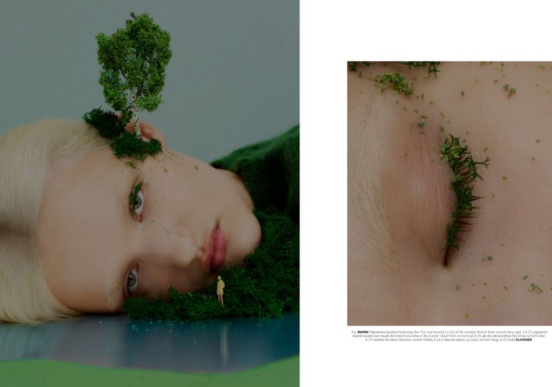 Close-up of a blonde model with miniature moss landscape and trees on her head and face against a grey studio backdrop.