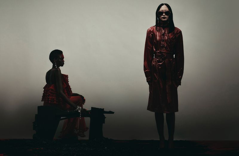 Two models in red fashion apparel against a muted gradient background, one standing and one seated.
