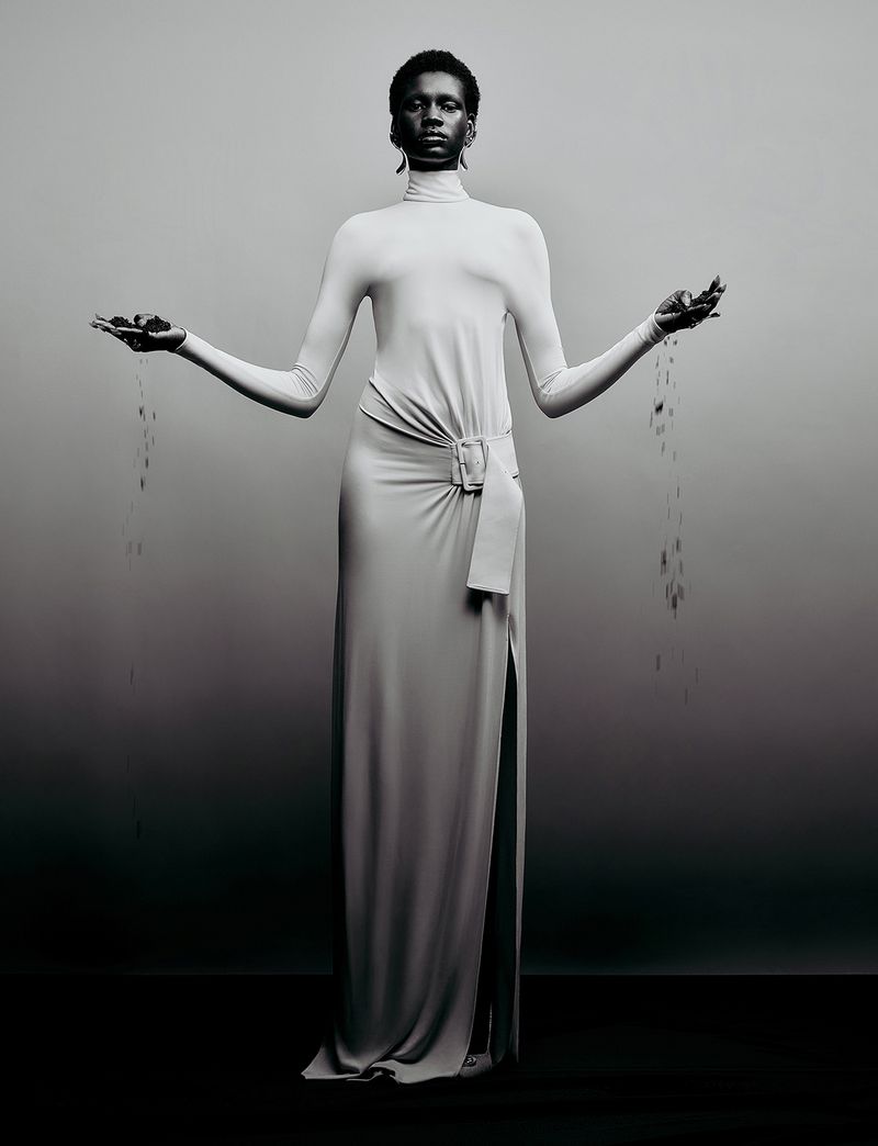 Model Nyagoa Diu wearing a high neck cream dress holding dark particles falling from hands against a grey studio backdrop