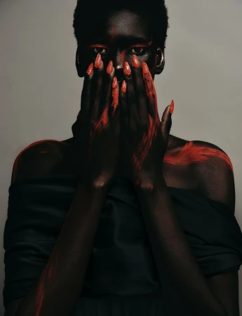 Close-up portrait of a Black model with hands held up covering the lower face, featuring striking red nail polish and crimson markings against skin and hair.