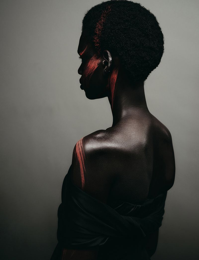 Profile portrait of a Black woman model with red face paint and hair accents against a neutral grey studio background.